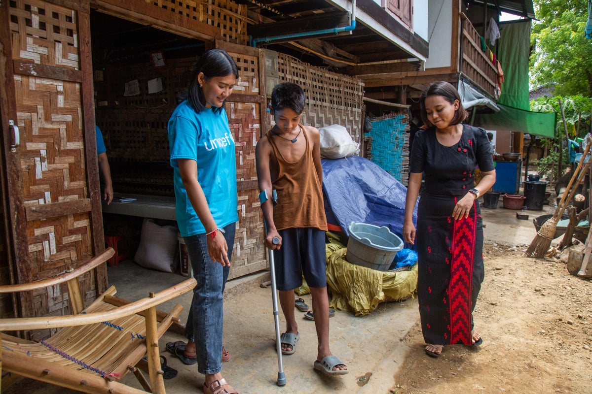 A boy uses a pair of forearm crutches to walk outside his home in Myanmar as his mother and a UNICEF worker watch. 