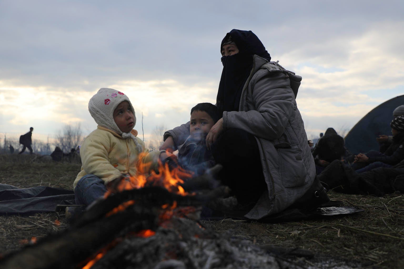 A woman and two children are seating on the floor by the fire.