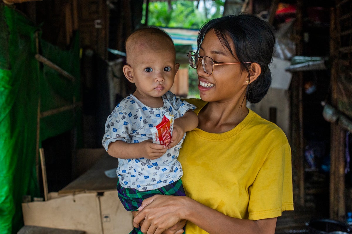 Hawng Nan holds her 20-month-old son, Hkawng Lum, in front of their shelter in a displacement site in Myanmar. Hkawng Lum holds a sachet of Ready-to-Use Therapeutic Food (RUTF).