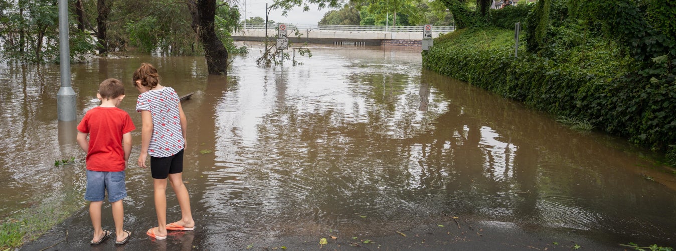 Two young children stand in front of floodwaters over a road.