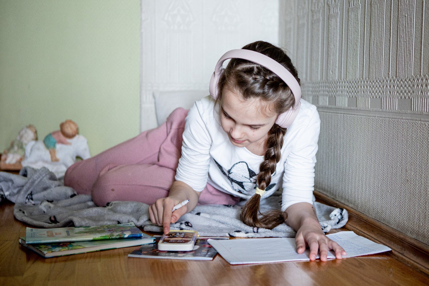 A 9-year-old-girl wearing pink headphones, pink pants and a white long sleeve top plays with her phone on her bedroom floor. 