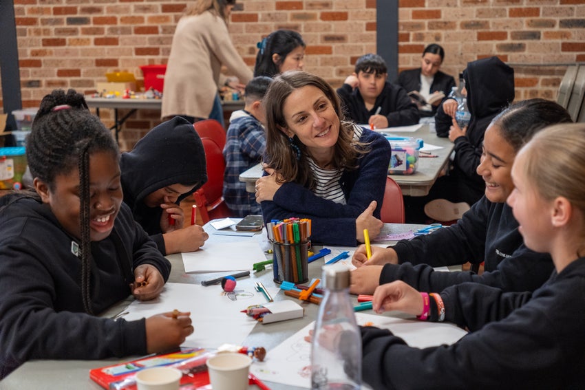 A group of children sit around a table completing activities as they chat with an adult facilitator. 
