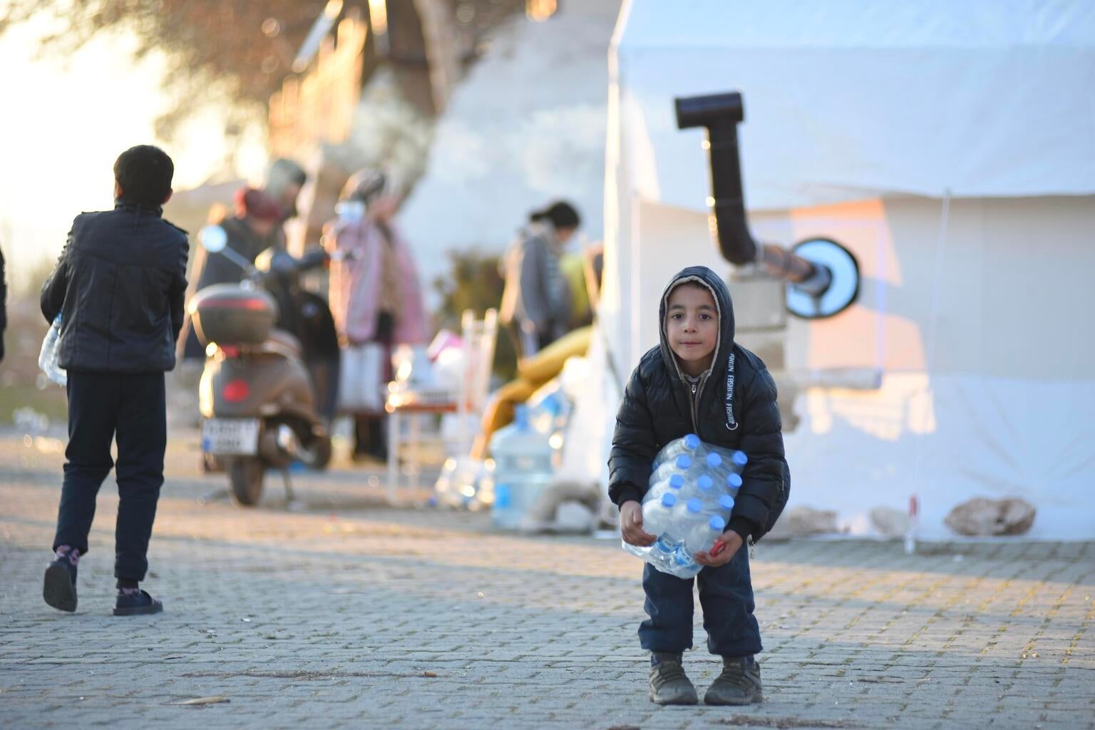 A little boy trying to carry a heavy water pack in a temporary shelter.