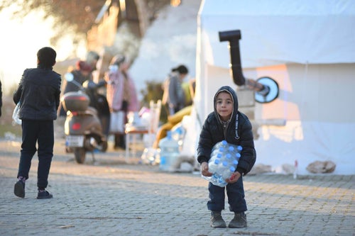 A little boy trying to carry a heavy water pack in a temporary shelter.