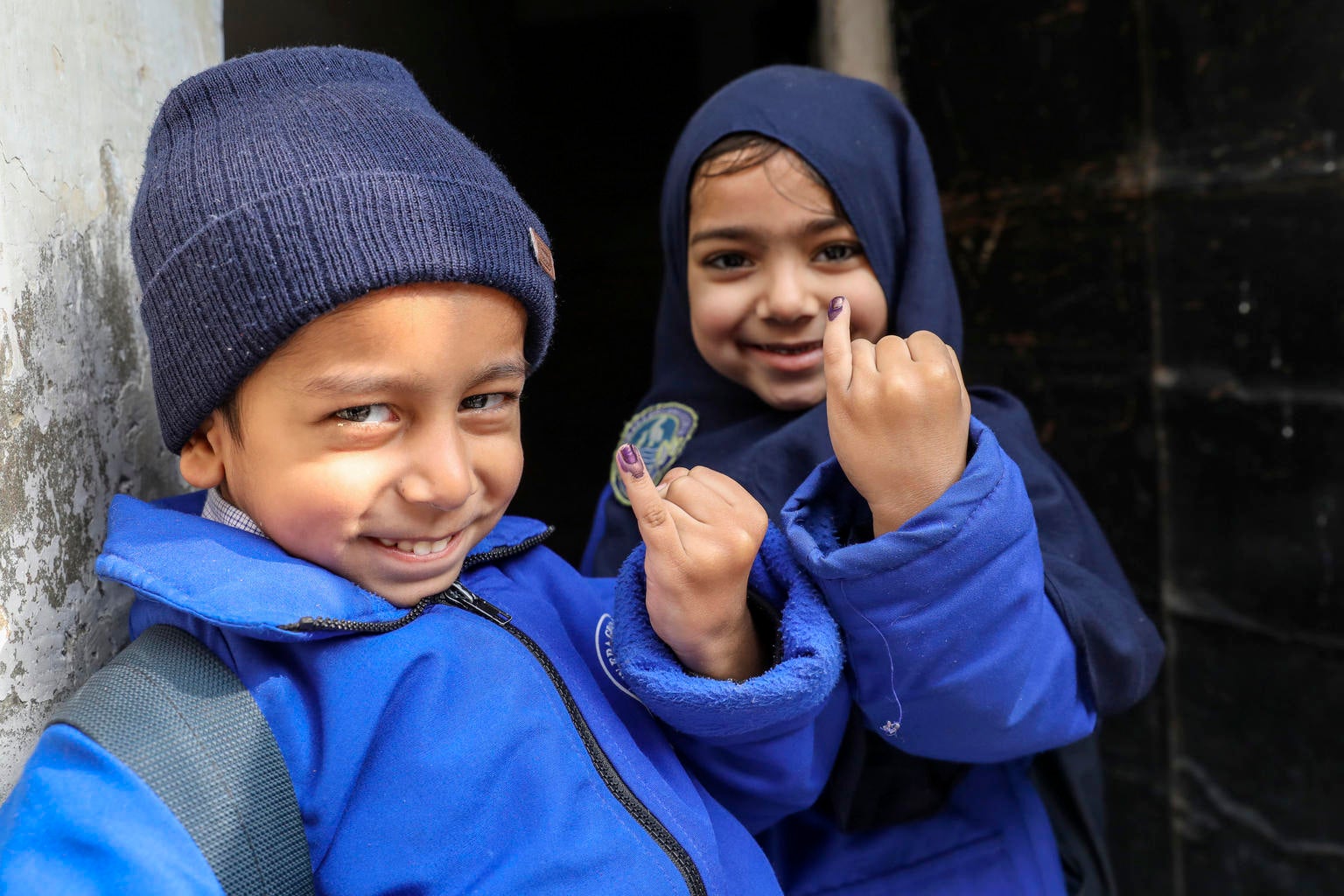 Two children in blue hold their pinky fingers out to show their vaccination mark.
