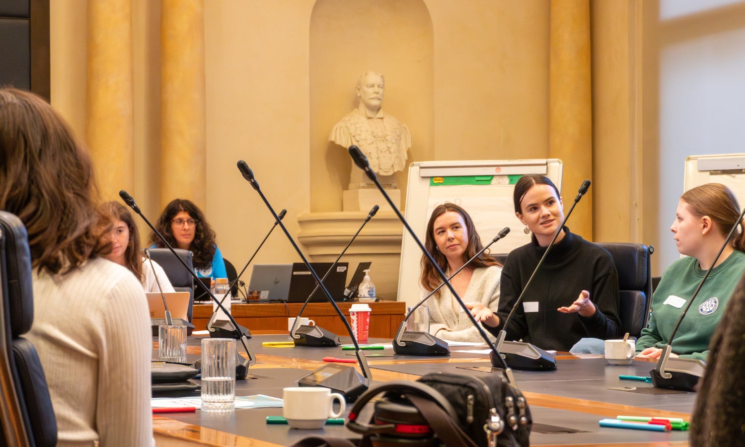 A group of young people seated around a table in the City of Sydney council chambers look on as a young woman speaks.