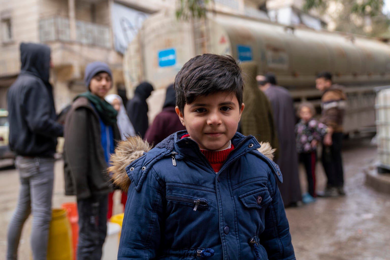 A young Syrian boy wearing a blue parka stands in front of a UNICEF water truck.