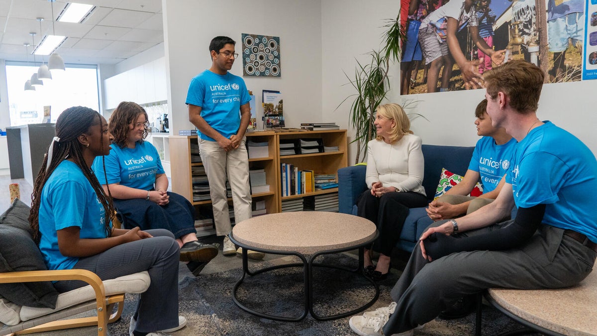 A woman in a white top talks to a group of young adults in blue tshirts. 