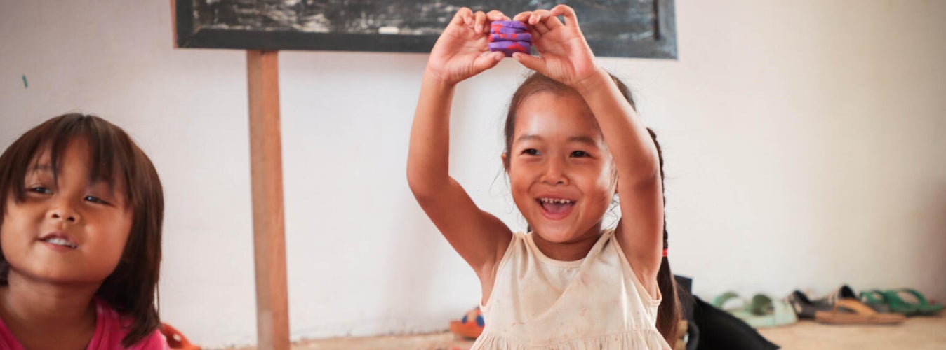 Two smiling young girls sitting on a rug in a classroom in Laos.  