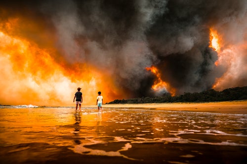 Two children stand in the shallows on a beach as a bushfire burns in the distance