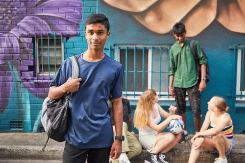 A teenage boy with a backpack stands in the foreground as a group of young people talk on a city sidewalk in the background.  