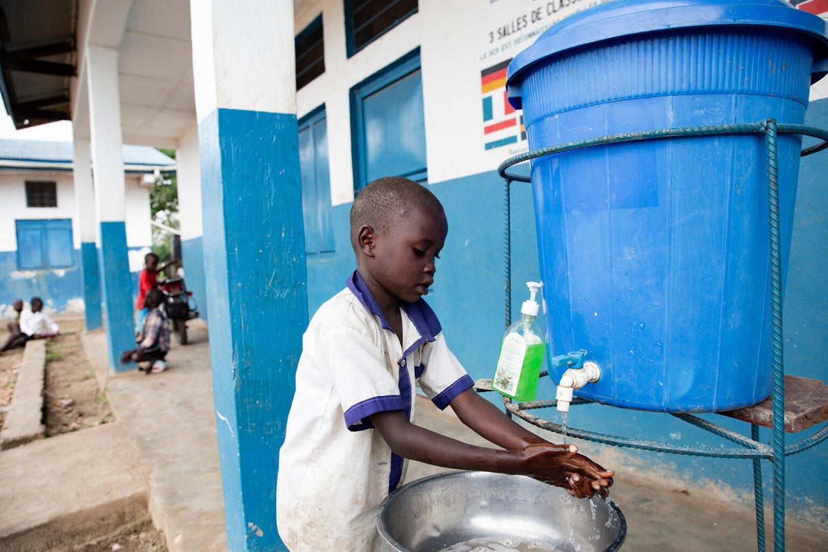 School boy washes hands from large blue bucket with tap.
