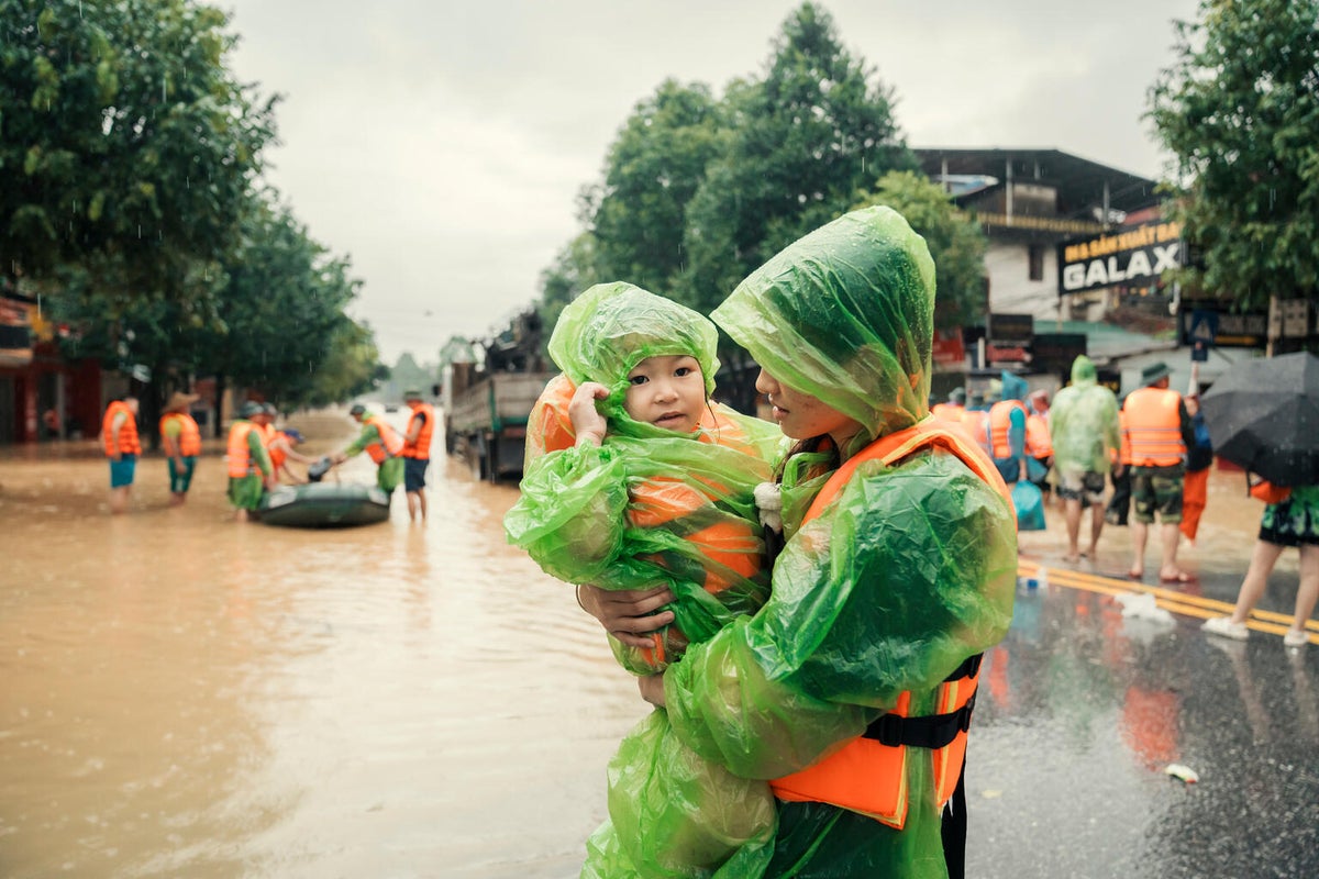 A teenage girl in a raincoat carrries her younger sister in a flooded street.