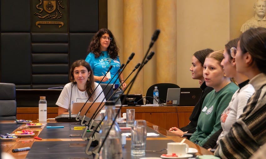 A group of young people seated around a table in the City of Sydney council chambers look on as a young woman speaks.