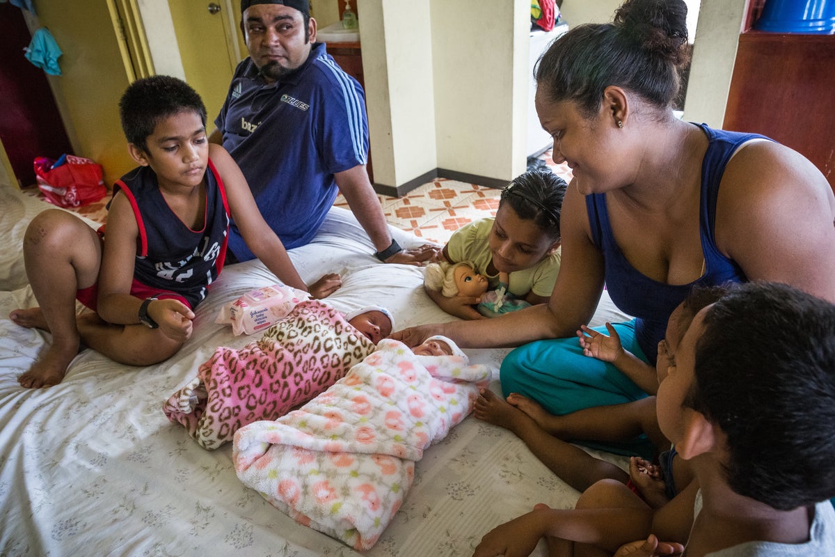 A man, a woman and three children seat around a double bed. The two newborns are lying in the middle of the bed.