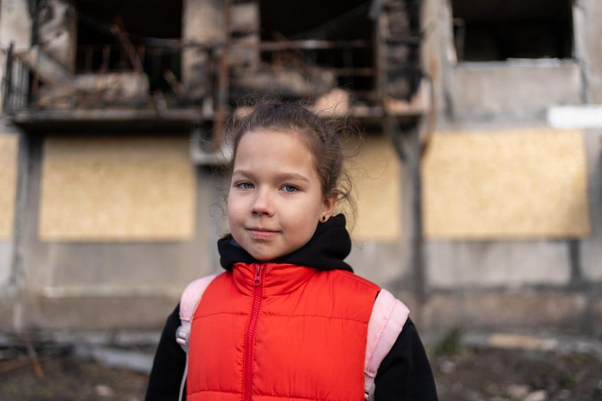 In Ukraine, eight-year-old Myroslava stands in front of a bombed-out apartment block, one of several damaged in a missile strike