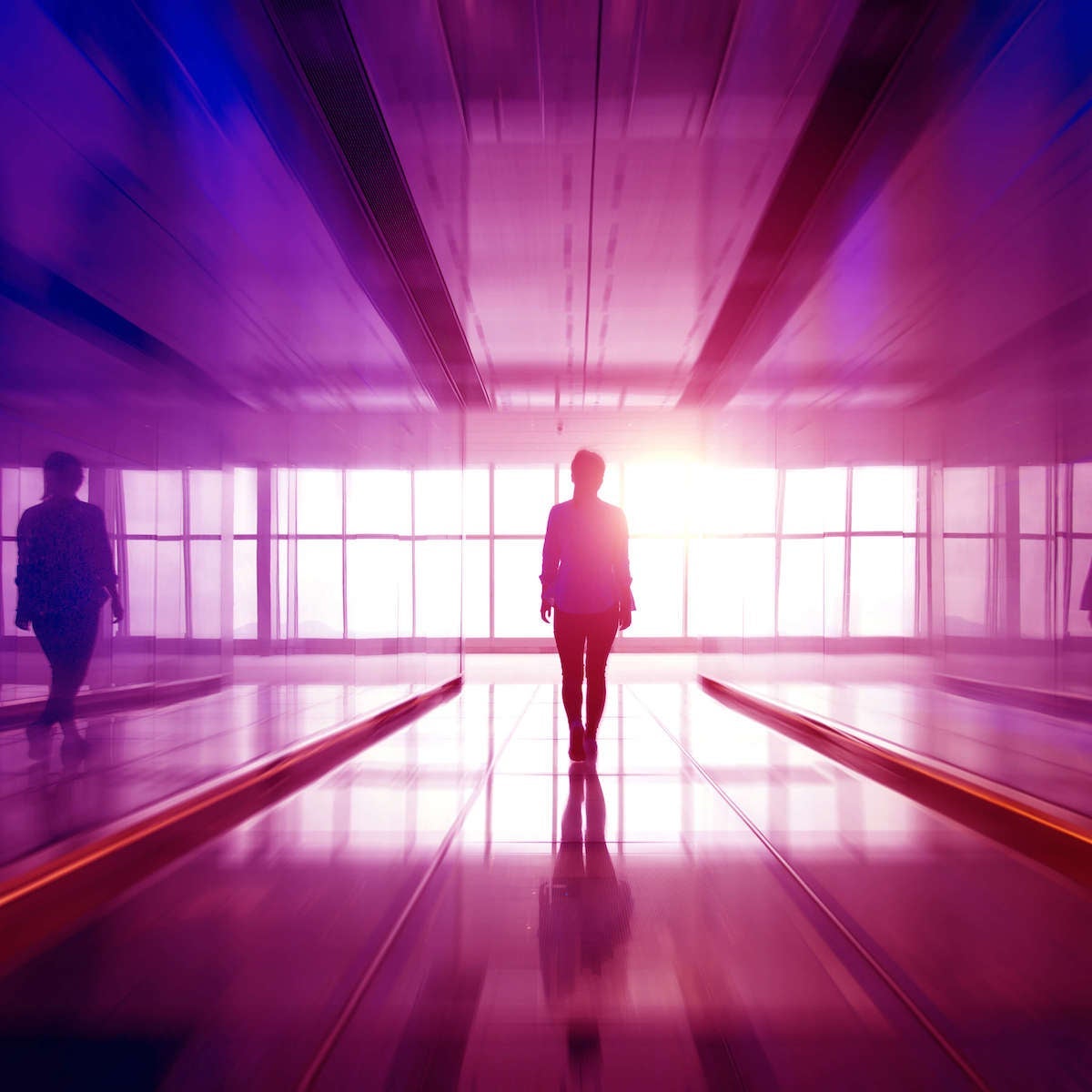 A person walks down a hallway with glass walls toward a set of floor-to-ceiling glass windows as the sun rises