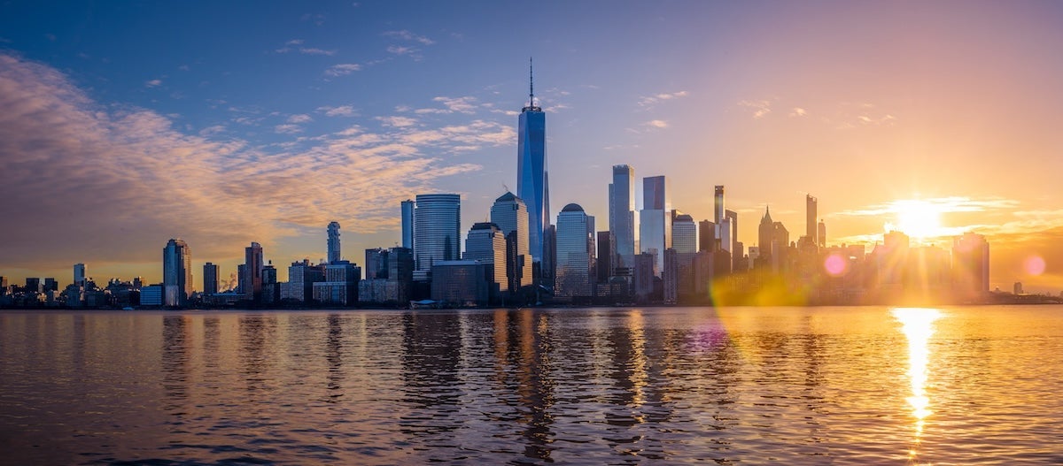 The sun rises above the New York City skyline and reflects off the water.