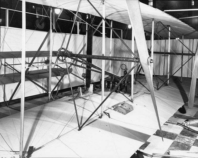 Engine built by Charlie Taylor in place on the wing of the Flyer; the pilot (Wilbur or Orville) would lie on the cradle next to the engine, balancing its weight (airandspace.si.edu)