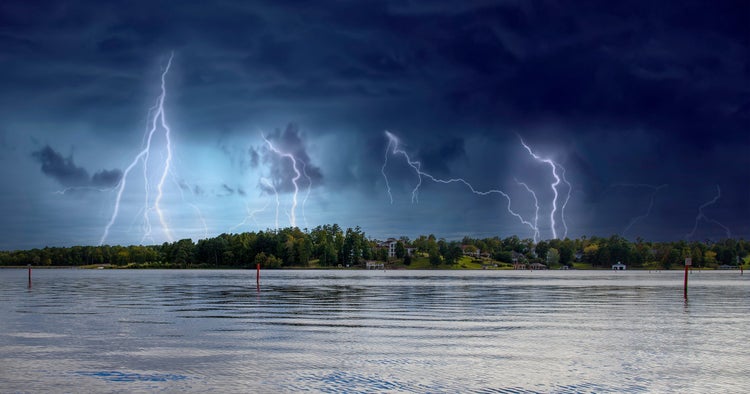 A dramatic storm scene of multiple lightning strikes illuminating a dark, stormy sky over a calm body of water, with trees and houses along a shoreline.