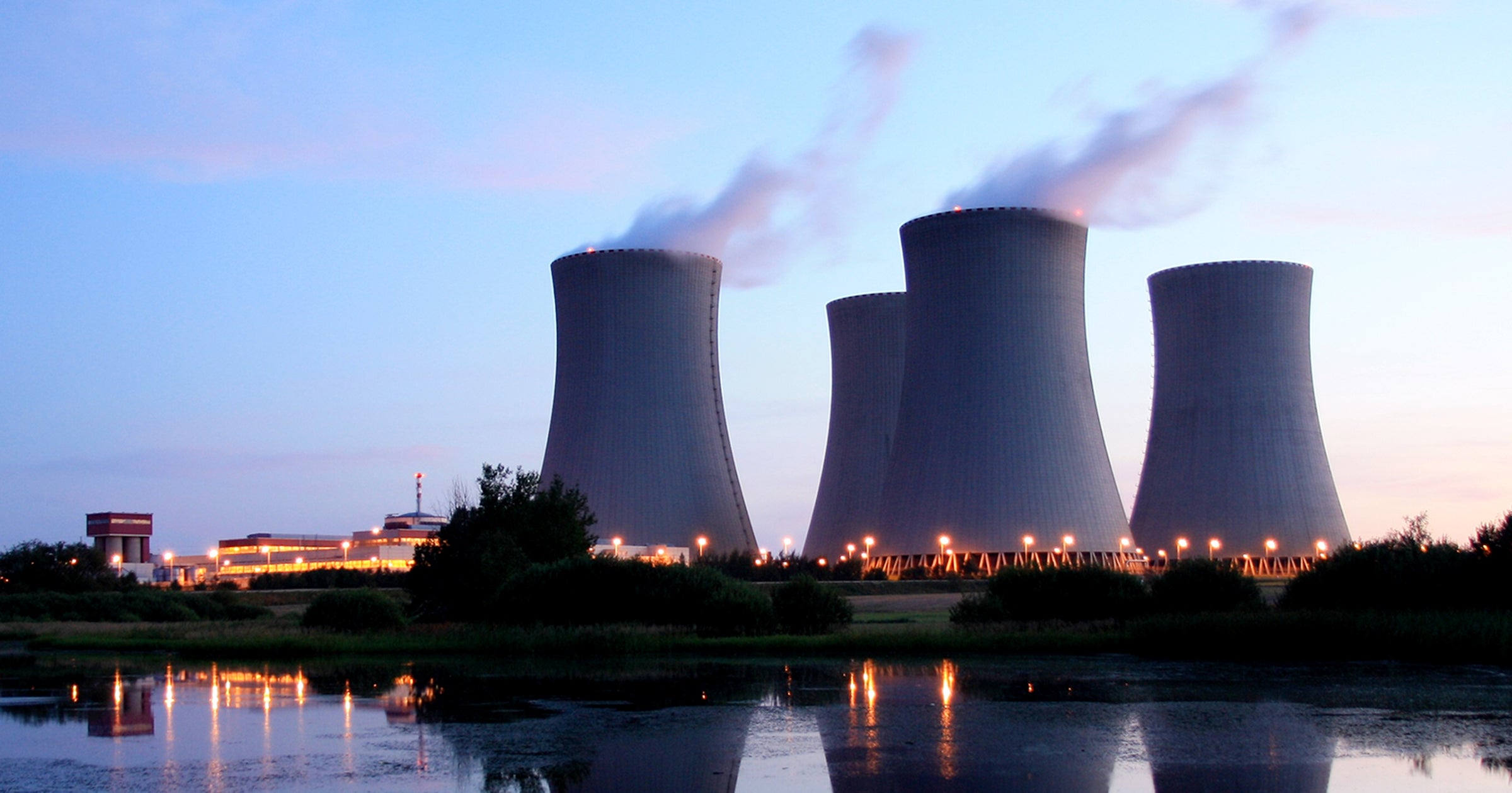 A power plant with multiple large cooling towers emitting steam, illuminated at dusk with reflections in the water.