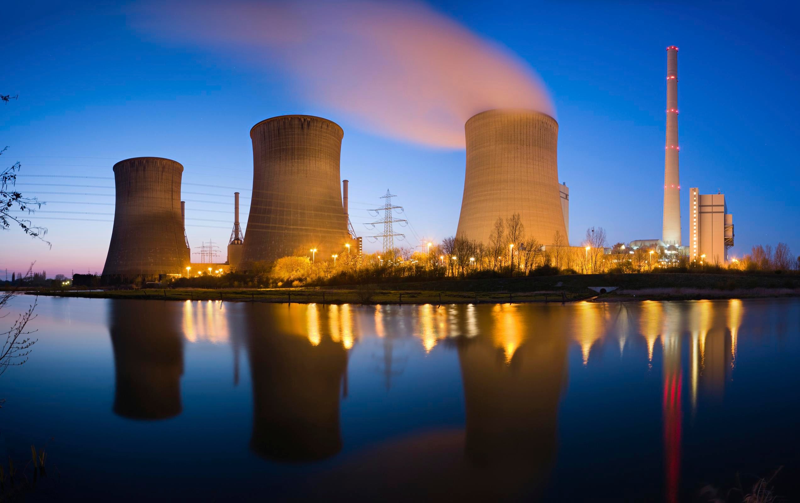 A power plant with four cooling towers emitting steam and a tall chimney, all reflected on a calm body of water during twilight