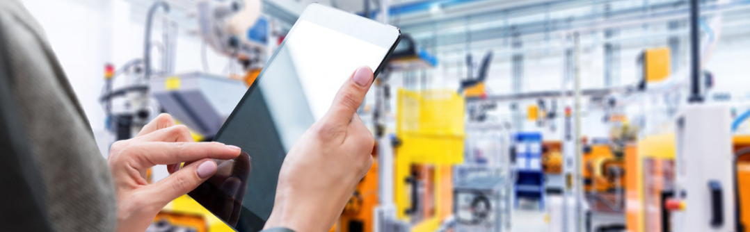 Horizontal color image of businesswoman - unrecognizable person - working with digital tablet in large futuristic factory. Woman standing on top of a balcony, holding touchpad and checking inventory of a manufacturing company on touchscreen tablet. Focus on businesswoman's hands holding black tablet, futuristic machines in background.