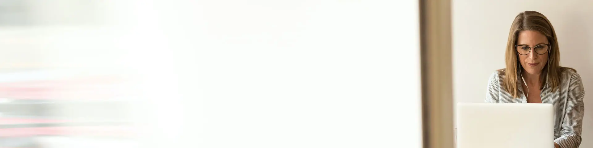 A middle-aged woman working on her laptop while listening to music near a window.