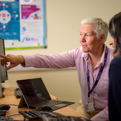Two employees sit at a computer monitor. A woman in pink is pointing at the screen. 