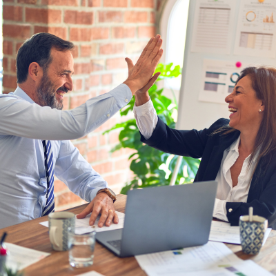 A man and a women high five while sitting next to a laptop and sheets of paper. 