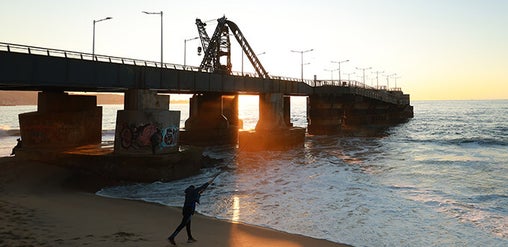 Muelle industrial al atardecer, con grúa, pilares con grafitis y olas del Pacífico; persona camina por la playa en Valparaíso, Chile.