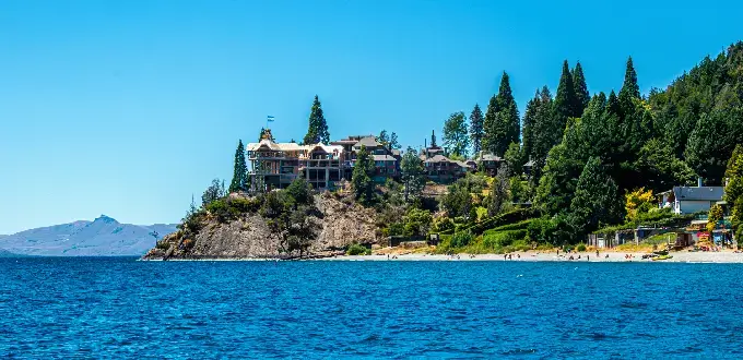 Costa del lago Nahuel Huapi en Bariloche, con playa, bosque patagónico y montañas al fondo.