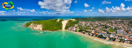 Vista panorámica de la playa de Ponta Negra en Natal, Brasil, con mar turquesa, arena dorada, dunas verdes y ciudad costera bajo cielo azul con nubes.