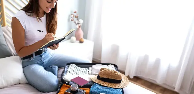 Mujer preparando maleta y planificando viaje en dormitorio, con ropa, sombrero y pasaporte listos para salir.