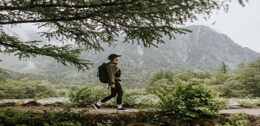Mujer con mochila haciendo senderismo por un sendero natural rodeado de montañas y bosque frondoso.