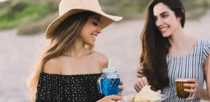 Mujeres disfrutando un picnic junto a la playa con bebidas refrescantes, ideal para vacaciones de verano.