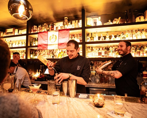 Dos bartenders preparando cócteles en la barra del Backroom Bar, frente a estantes iluminados con botellas y la bandera de Perú colgada como decoración.