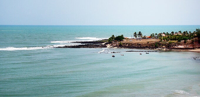 Costa tranquila en Baía dos Golfinhos, Pipa (Brasil), con aguas verdes, acantilados, palmeras y pequeñas embarcaciones cerca de la orilla.