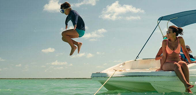Niño saltando al mar desde una lancha en Maragogi, Brasil, mientras su familia disfruta el día en aguas turquesa y cielo despejado.