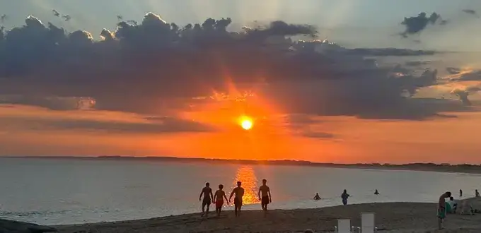 Atardecer en la playa de José Ignacio, Uruguay, con el sol sobre el mar, cielo anaranjado y personas caminando por la costa.