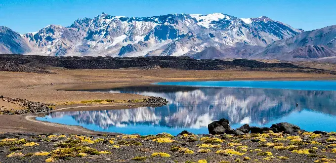 Lago rodeado de terreno árido con vegetación baja, reflejando montañas nevadas de la cordillera en Mendoza, Argentina.