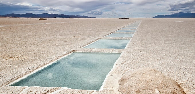 Superficie blanca de Salinas Grandes en Salta-Jujuy, con piletones de agua turquesa y montañas en el horizonte bajo cielo nublado.