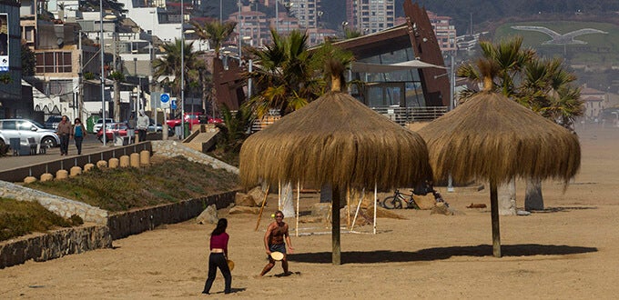 Personas caminan y juegan en la arena junto a palapas en Playa Reñaca, balneario urbano de Viña del Mar, Chile.