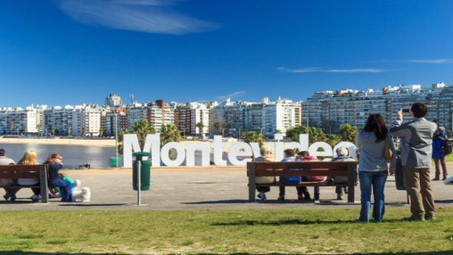 Turistas fotografiándose frente al icónico cartel de Montevideo en la rambla, con edificios al fondo y cielo despejado.