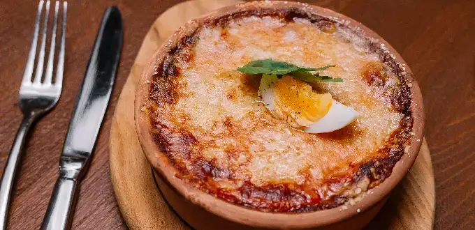 Pastel de choclo chileno en fuente de greda, gratinado, con huevo duro y albahaca sobre mesa de madera.