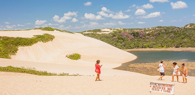 Dunas blancas y laguna de Genipabu en Natal, Brasil, con visitantes caminando entre arena, vegetación costera y cielo despejado.