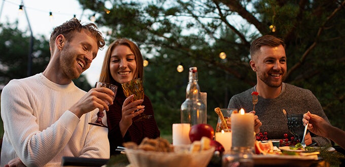 Grupo de amigos disfruta una cena al aire libre con velas, vinos y platos gourmet, ambiente cálido y social en jardín nocturno.