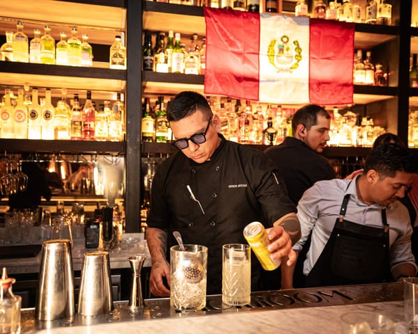 Bartender preparando un cóctel en Backroom Bar con la bandera de Perú al fondo y una amplia selección de licores en las estanterías iluminadas.