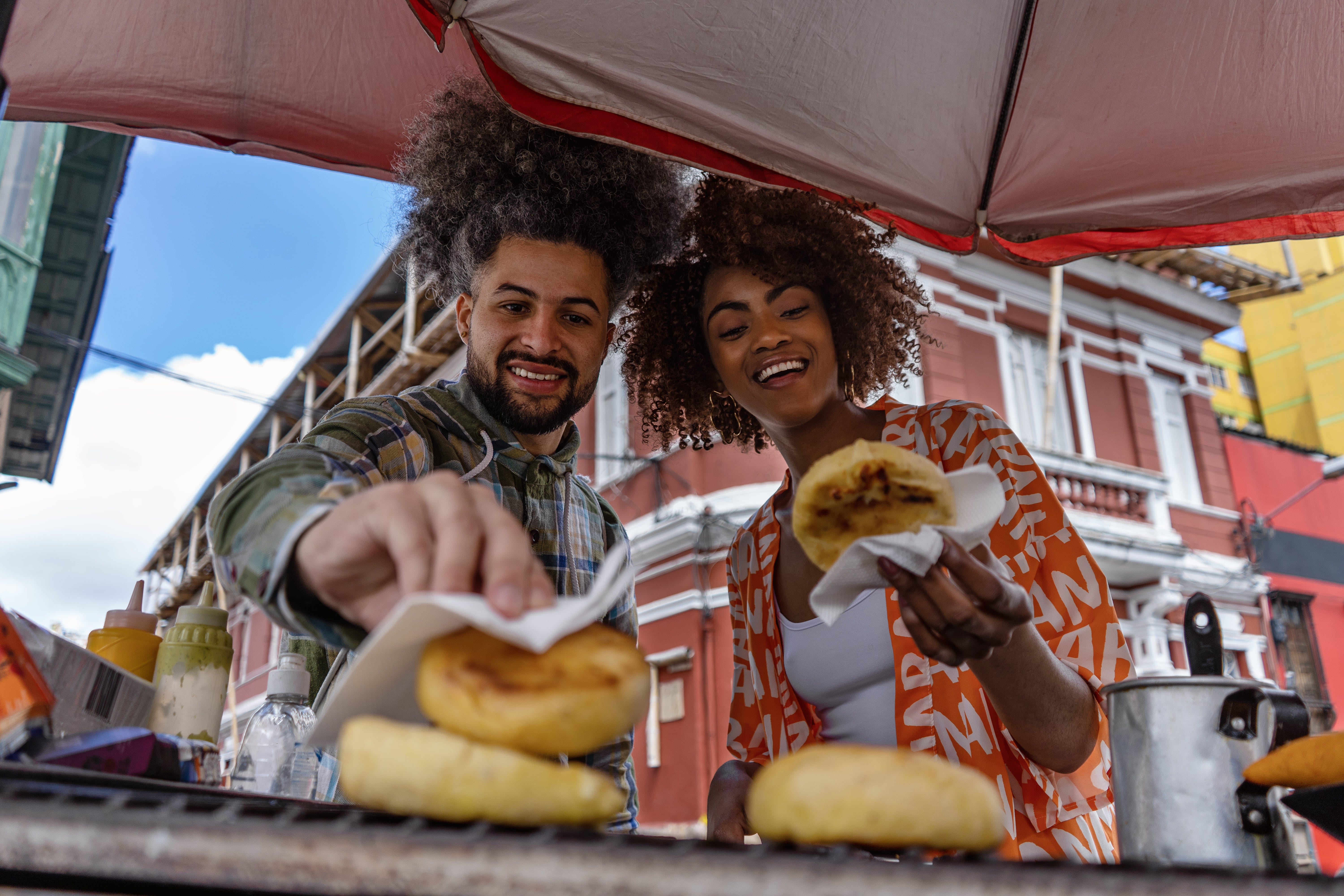 Pareja disfrutando arepas en un puesto callejero en Colombia, con casas coloniales de fondo.