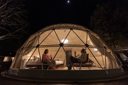 Pareja en domo glamping de noche en zona montañosa, con cielo despejado y paisaje abierto.
